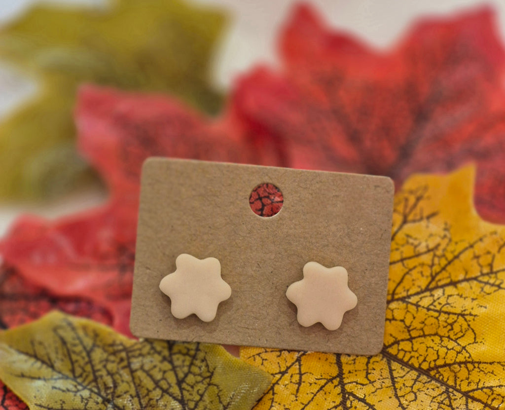 Nude polymer clay stud earrings shaped like six-petal flowers, displayed on a brown card surrounded by red, yellow, and green autumn leaves.