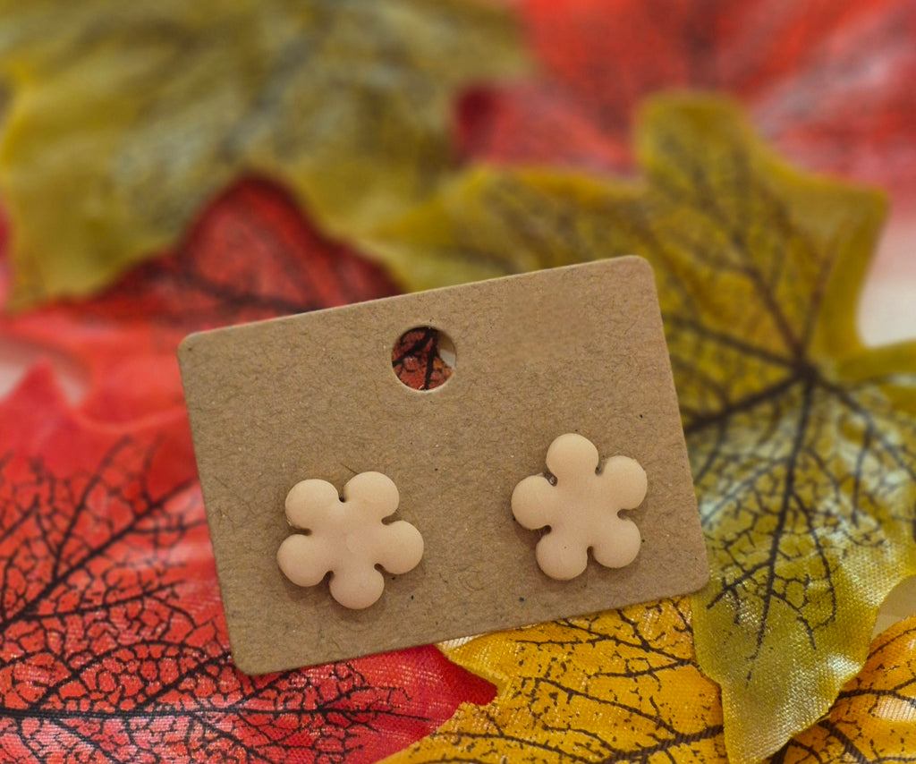 Nude-tone polymer clay stud earrings shaped like four-petal flowers, displayed on a brown backing card surrounded by red, yellow, and green autumn leaves.