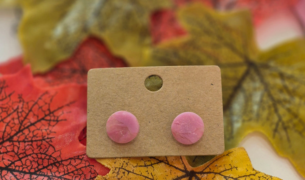 Pink marble-pattern polymer clay stud earrings displayed on a brown backing card surrounded by autumn leaves in red, yellow, and green tones.