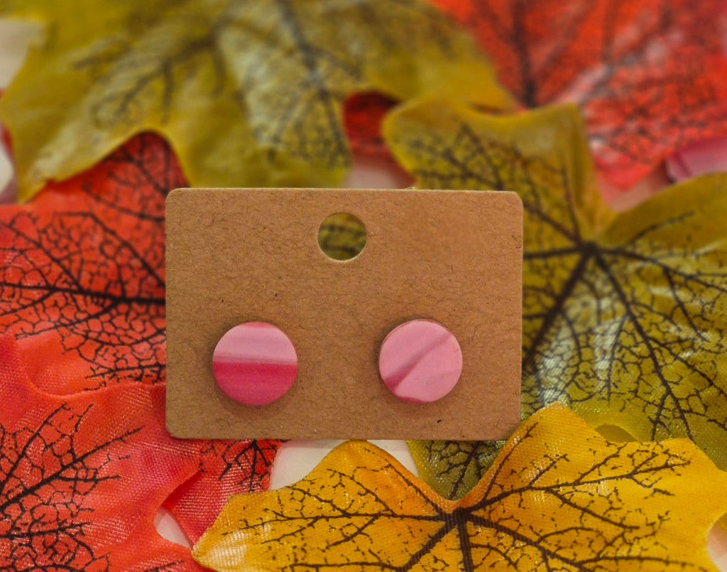 Pink marbled polymer clay stud earrings displayed on a brown backing card surrounded by colourful autumn leaves in red, yellow, and green.