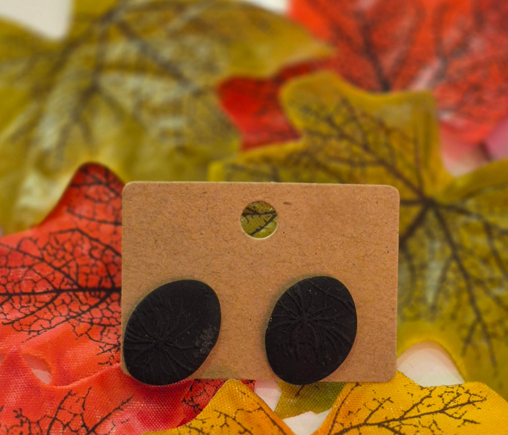 Matte black oval polymer clay earrings with subtle flower imprint design, displayed on a brown backing card surrounded by red, yellow, and green autumn leaves.
