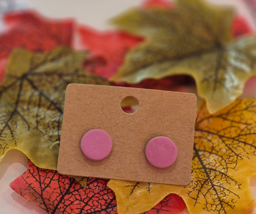 Light pink clay stud earrings displayed on a brown backing card surrounded by red and yellow autumn leaves.