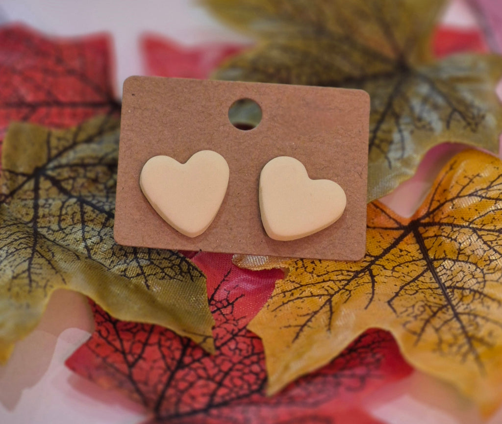 Cream heart-shaped clay stud earrings displayed on a brown backing card surrounded by autumn leaves in red, yellow, and green tones.