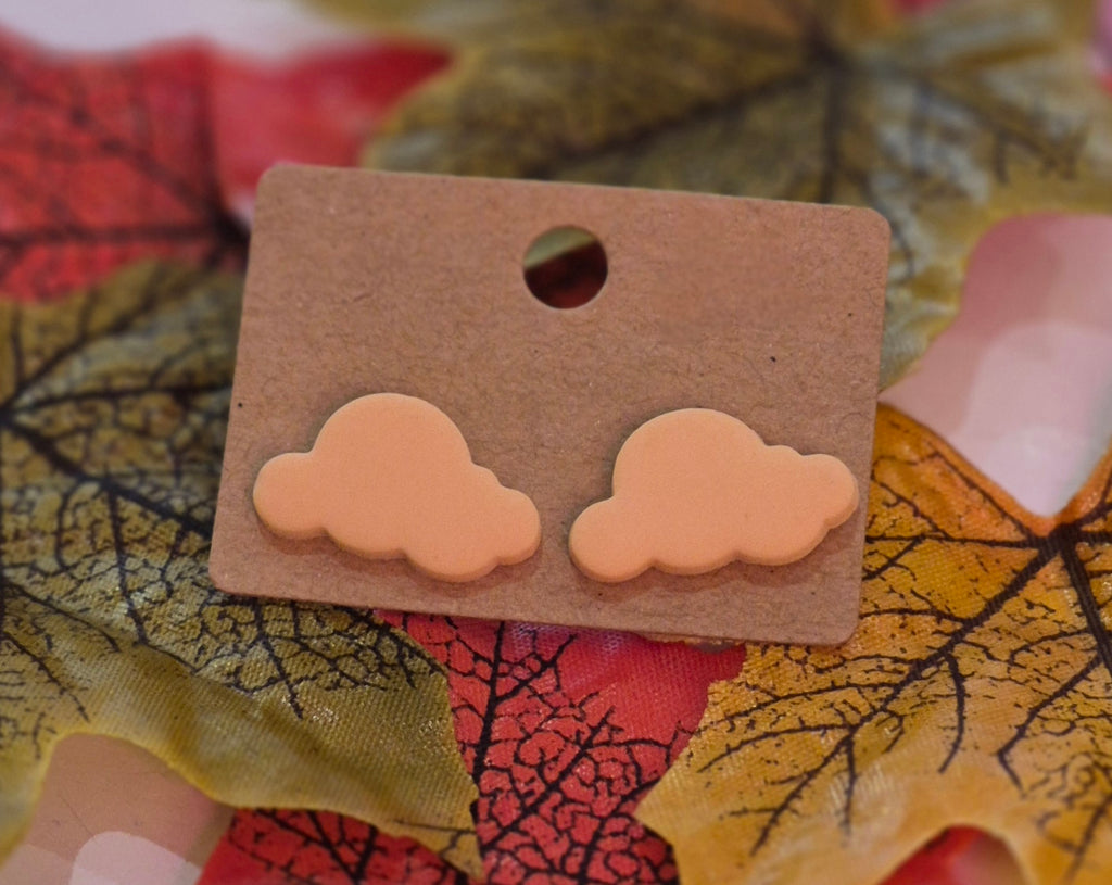 Peach-coloured cloud-shaped stud earrings displayed on a brown backing card surrounded by autumn leaves