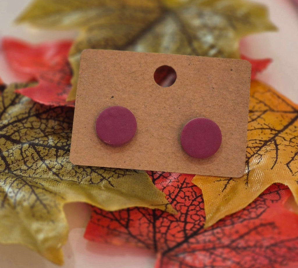 Deep berry purple round stud earrings displayed on a brown backing card surrounded by autumn leaves.