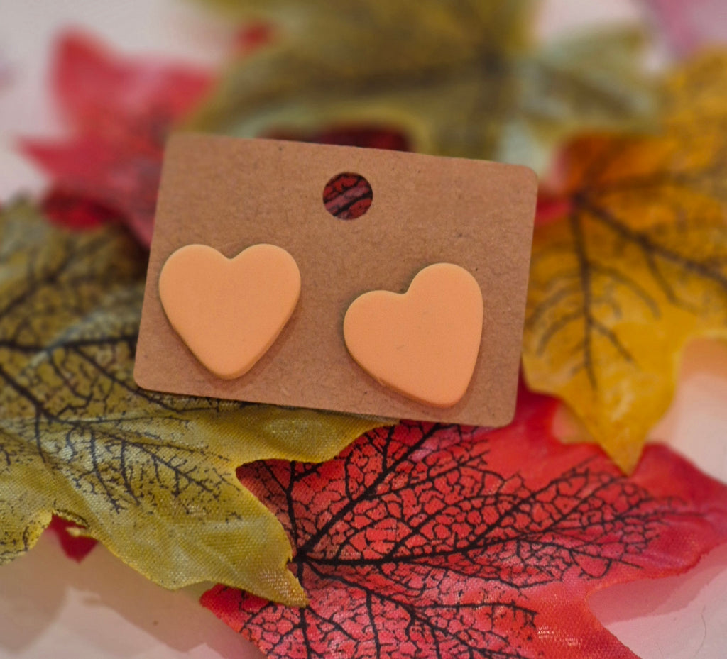 Peach heart-shaped clay stud earrings displayed on a brown backing card surrounded by yellow and red autumn leaves.