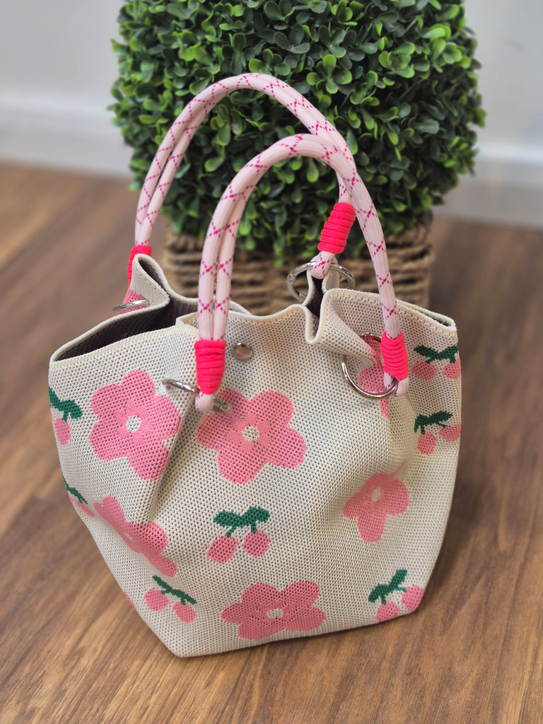 Cream tote bag with pink flowers and cherry designs, featuring thick rope handles with pink accents, placed on a wooden floor with a plant in the background.