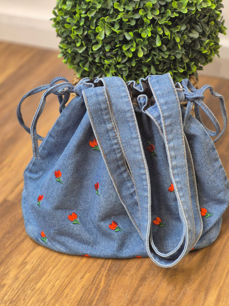 Denim tote bag with small red tulip embroidery, featuring drawstring closure and shoulder straps, placed on a wooden floor with a green plant in the background.