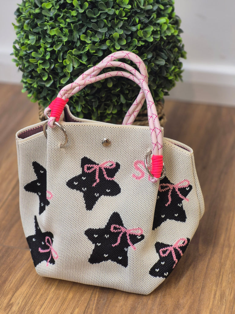 Cream tote bag with black star and pink bow pattern, pink rope handles, and silver hardware, placed on wooden floor with green plant background.