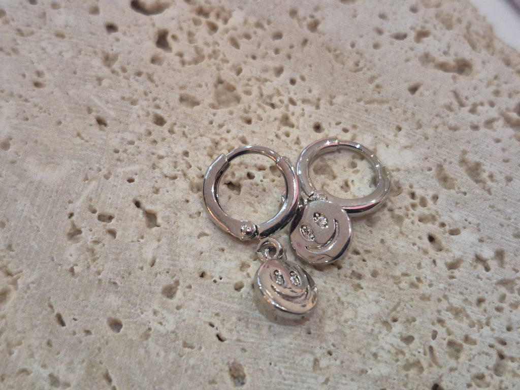 A pair of silver huggy hoop earrings with  smiley face charms displayed on a textured stone background.