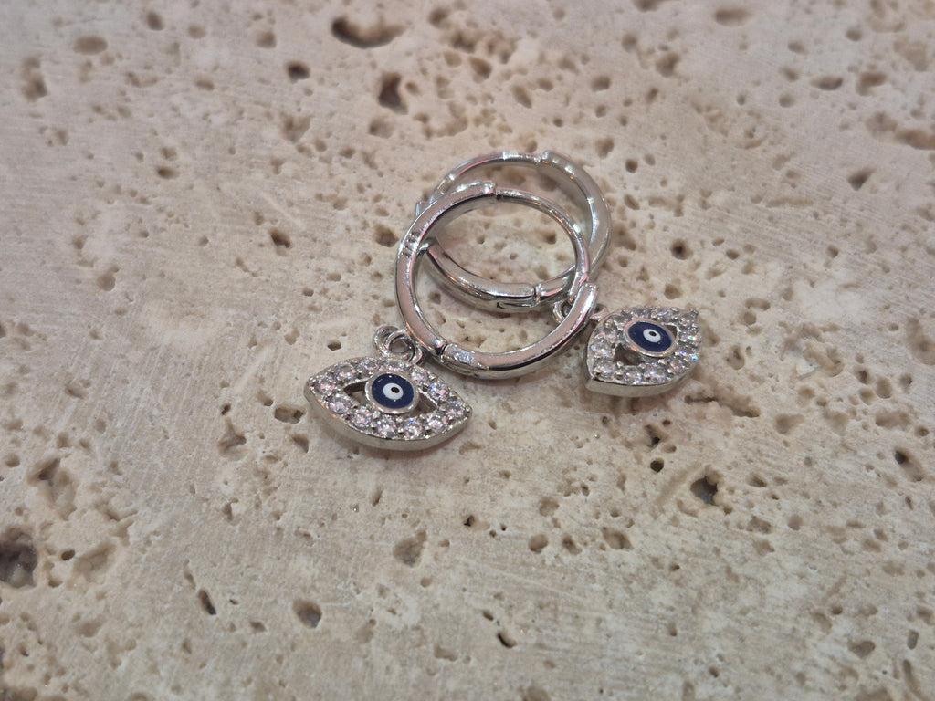 A pair of silver huggy hoop earrings with sparkling evil eye charms displayed on a stone background.