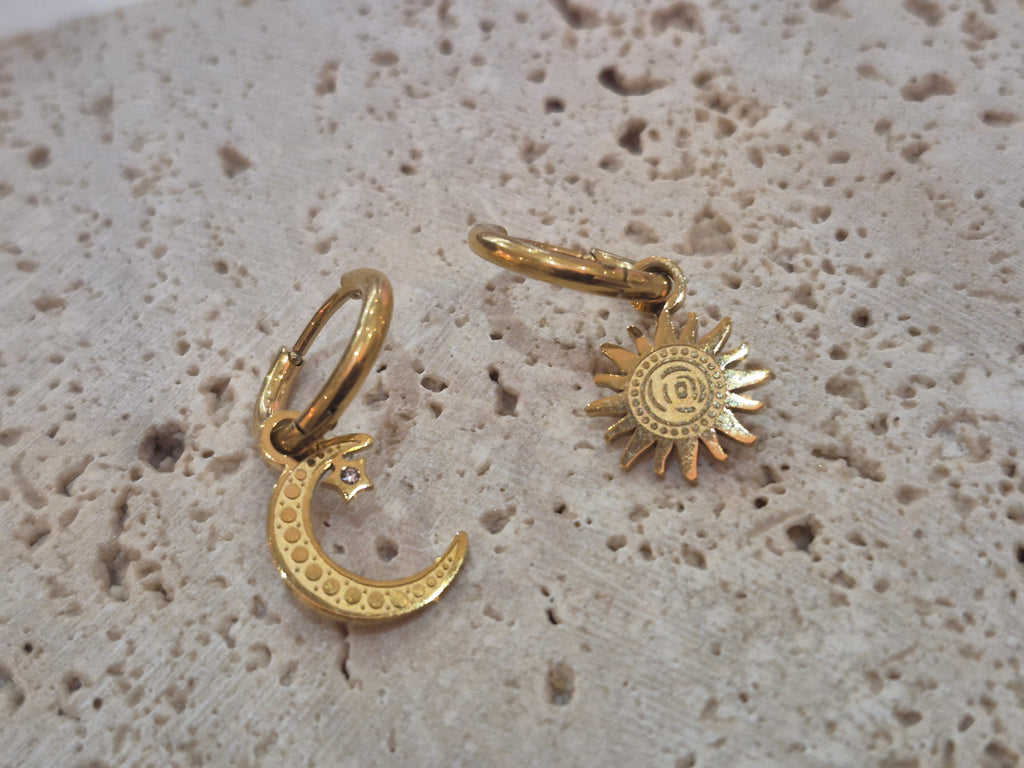 Gold-plated mismatched sun and moon huggy hoop earrings displayed on a textured stone background.