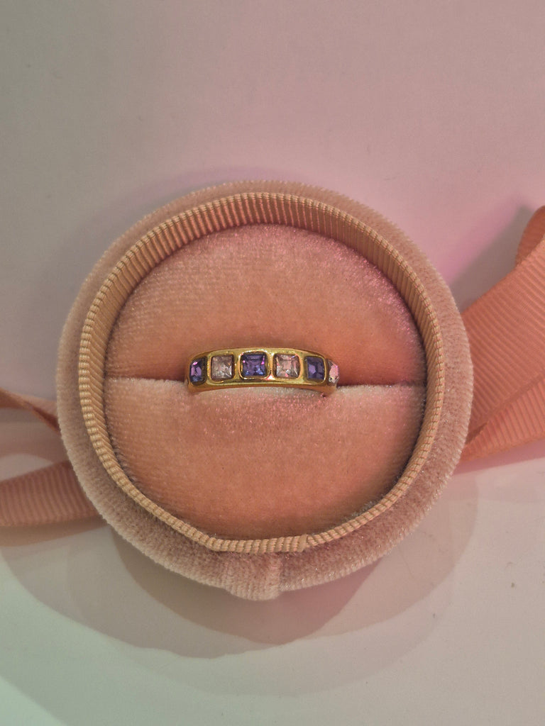 Gold band ring with alternating purple and clear gemstones displayed in a pink velvet ring box.