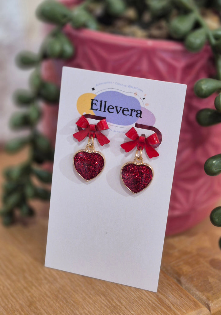 Red glitter heart earrings with small red bows displayed on Ellevera jewellery backing card.