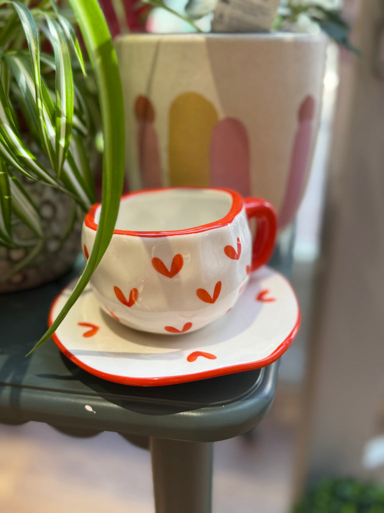 Ceramic cup and saucer with red heart patterns on a table with plants in the background