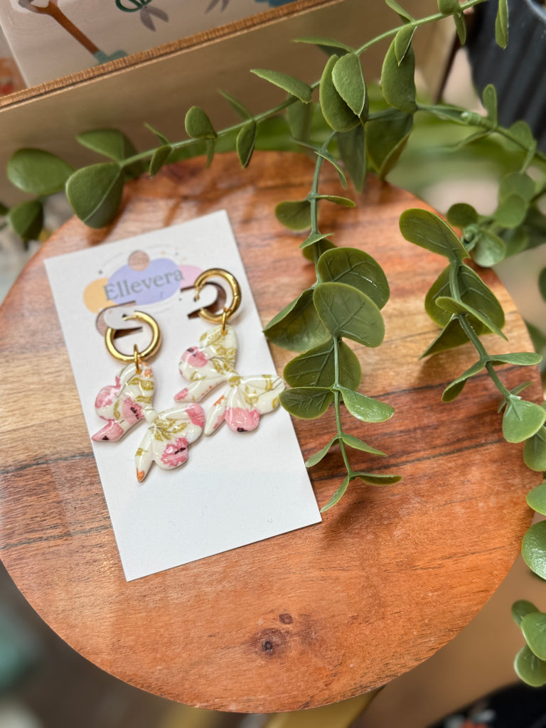 Handmade floral bow drop earrings with pink and gold accents on gold-tone huggie hoops, displayed on Ellevera backing card.