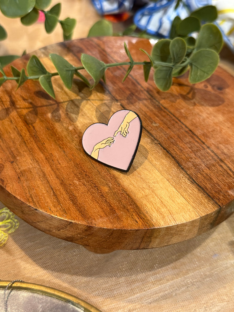 Pink heart enamel pin with two hands reaching towards each other