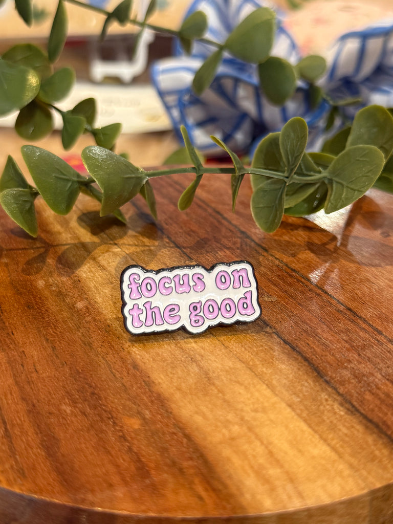 A pink enamel pin with the phrase “focus on the good” in bubble lettering, styled against a wooden background with green leaves.