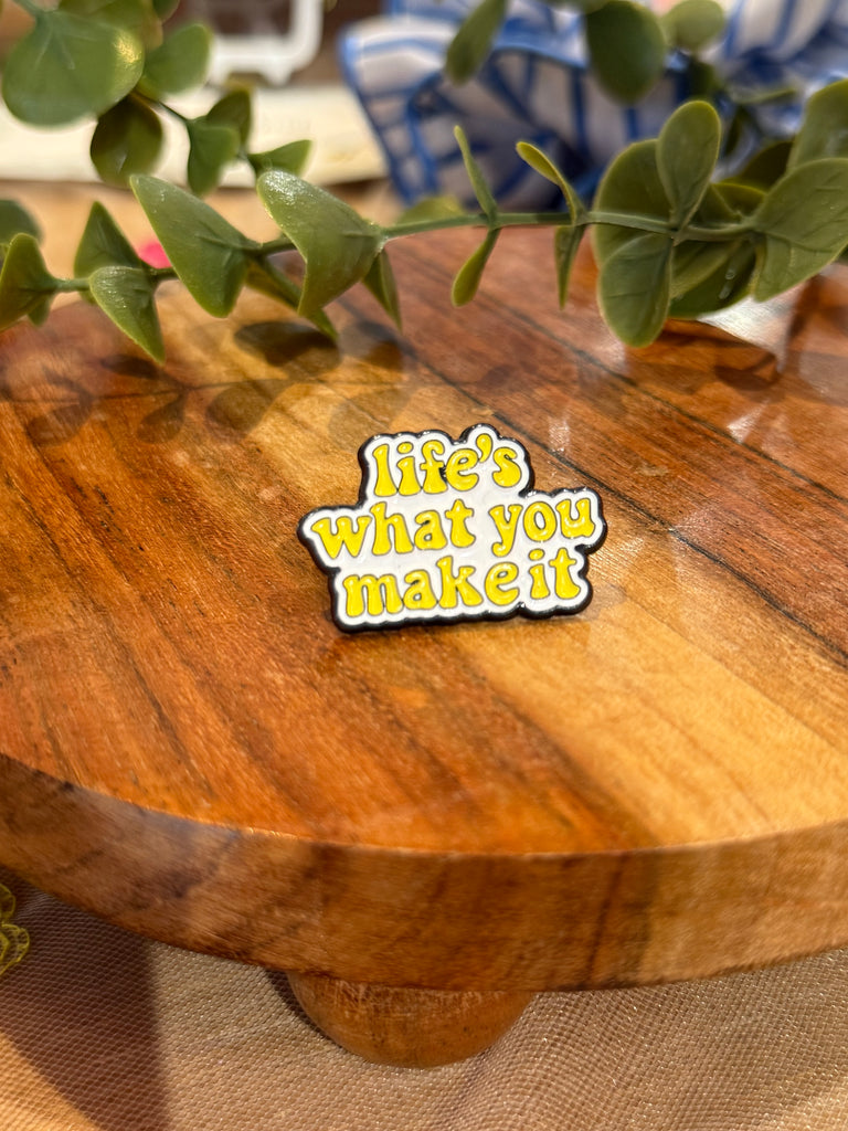 A yellow enamel pin with the phrase “life’s what you make it” displayed on a wooden surface with greenery in the background.