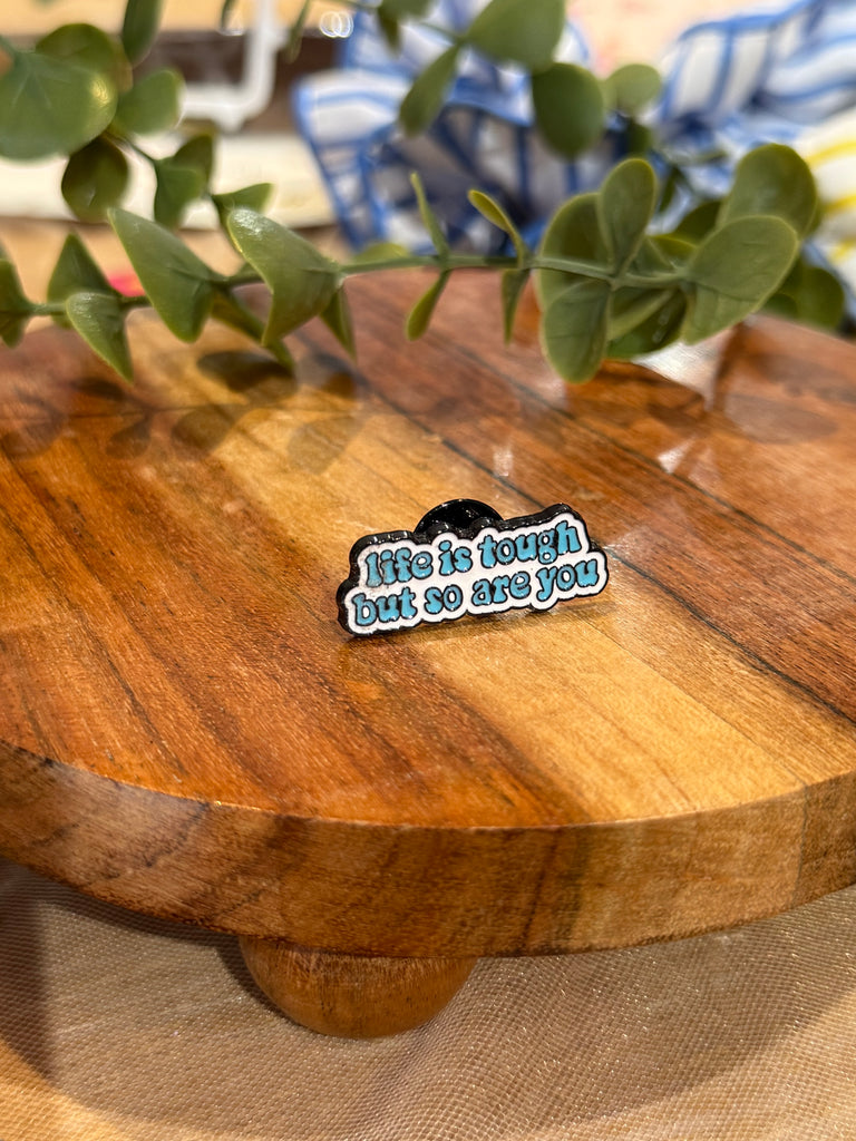 A blue enamel pin that reads “life is tough but so are you,” photographed on a round wooden board with green leaves in the background.