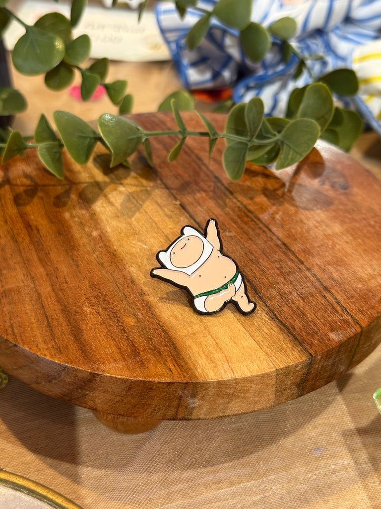Enamel pin of Baby Finn from Adventure Time in a diaper, smiling with arms raised, displayed on a wooden board with green leaves in the background.