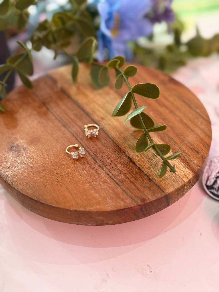 A gold bow crystal ear cuff with cubic zirconia detail displayed on a wooden stand with greenery.