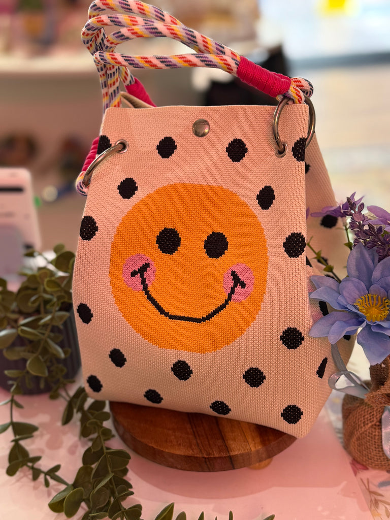 Beige woven tote bag with a large orange smiley face, black polka dots, and multicolour rope handles.
