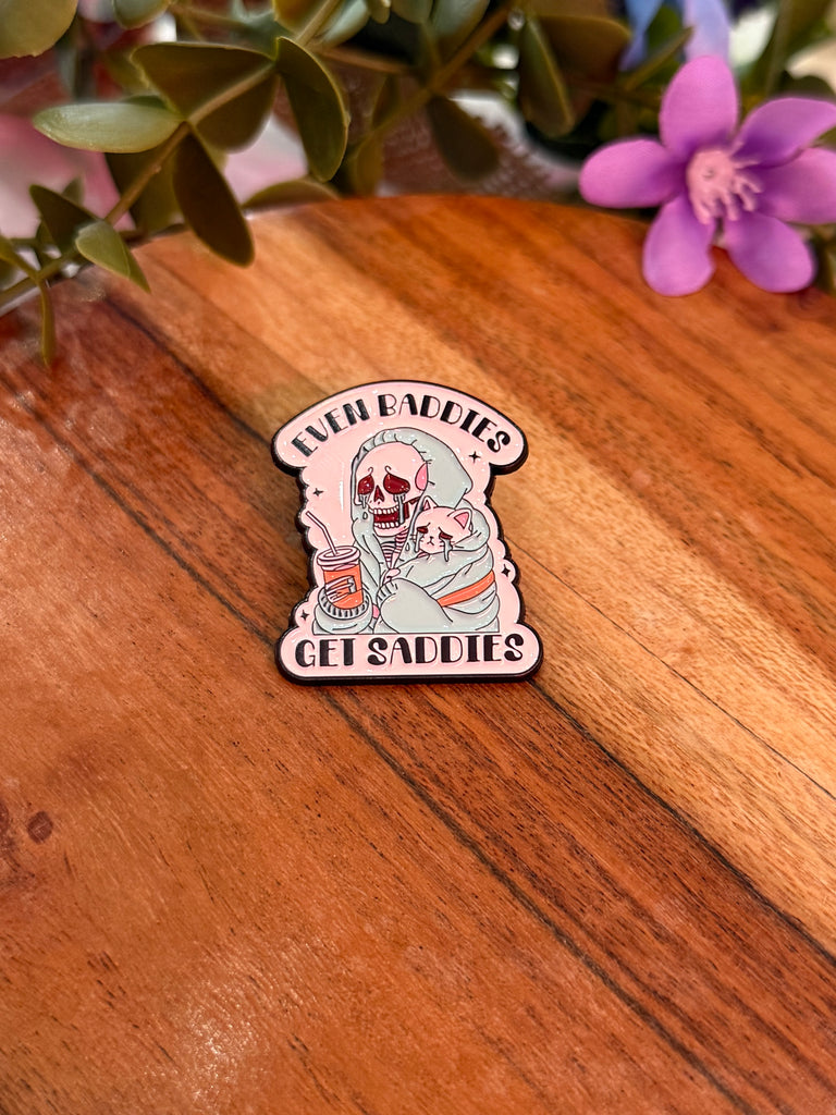 Enamel pin with skeleton holding a drink and text “Even Baddies Get Saddies.”