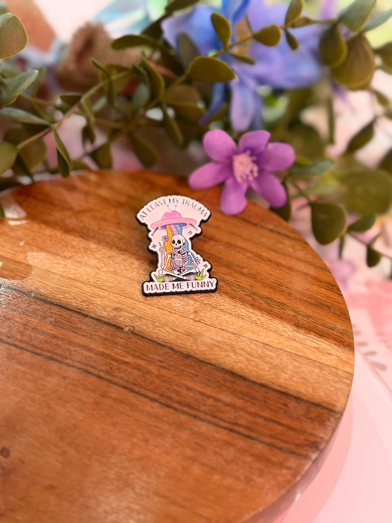 Enamel pin with skeleton holding an umbrella and text “At Least My Trauma Made Me Funny.”
