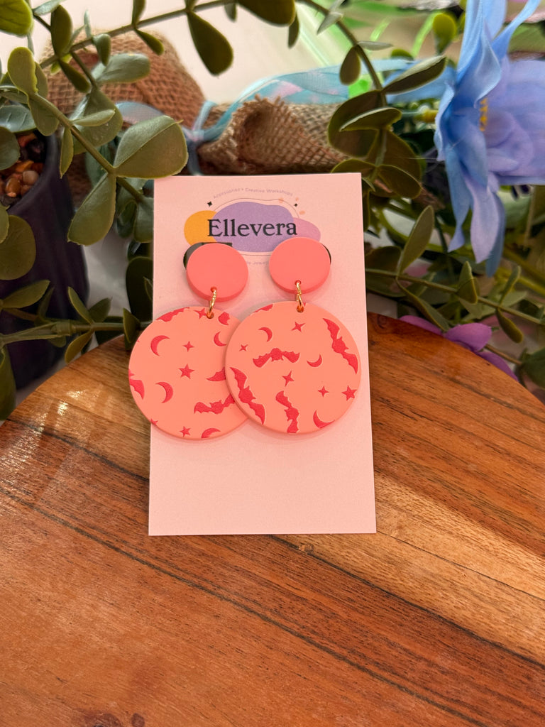 Round pink statement earrings with moon, star, and bat designs.