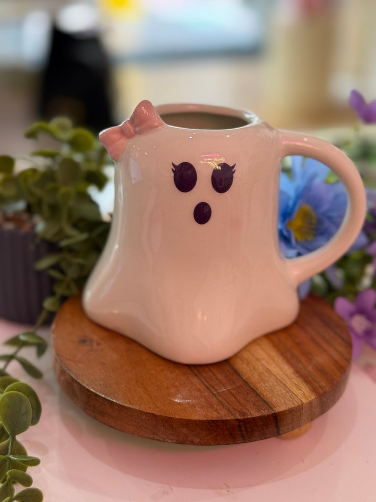 White ceramic mug shaped like a ghost with a pink bow and cute face, styled with greenery