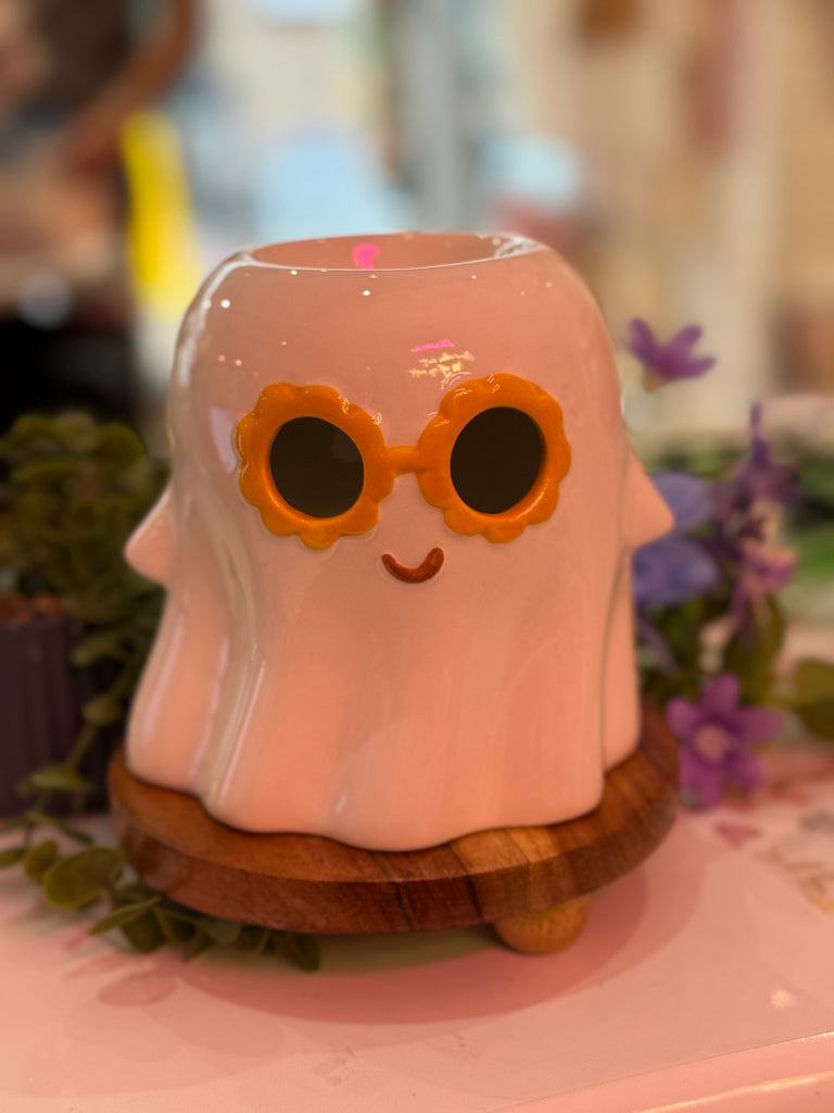 Ceramic ghost-shaped oil and wax melt burner with orange retro sunglasses, displayed on a wooden stand with flowers.