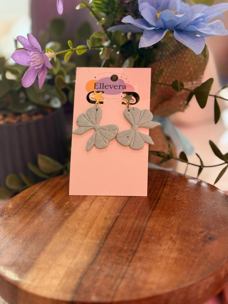 Blue polymer clay bow earrings with gold star studs, displayed on pink Ellevera card.