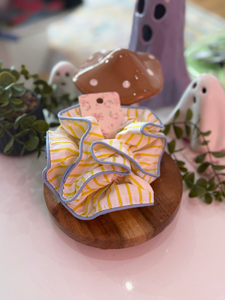 Yellow and white striped ruffle scrunchie with blue trim displayed on wooden stand.