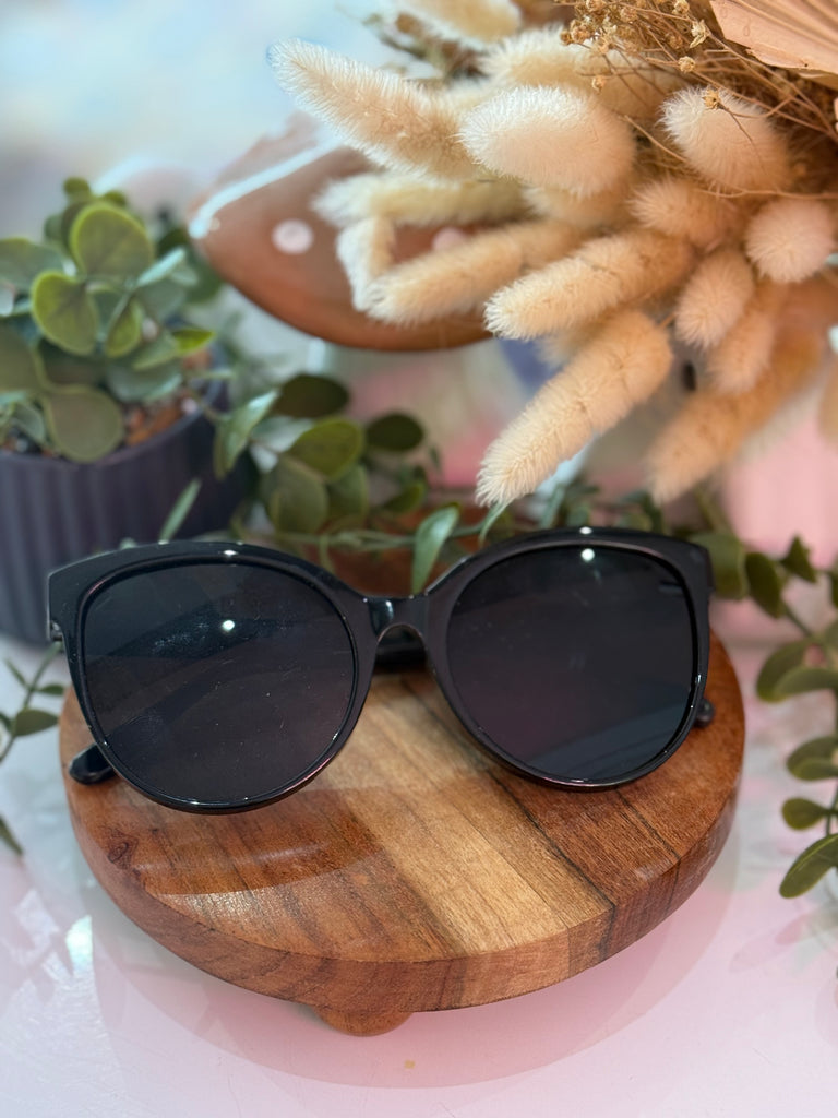 Black oversized cat-eye sunglasses with glossy frame displayed on wooden stand.