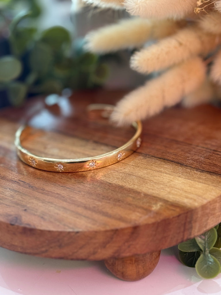 Golden sunburst bangle bracelet with engraved starburst details and crystals, displayed on wooden stand.