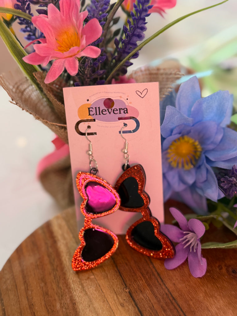 Red glitter heart sunglasses earrings displayed on Ellevera backing card with floral background.