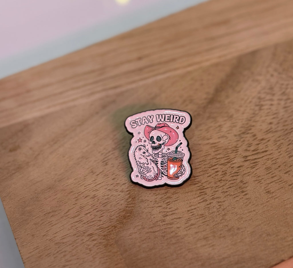Enamel pin with skeleton in a pink cowboy hat holding a drink and lizard, with the words "Stay Weird."