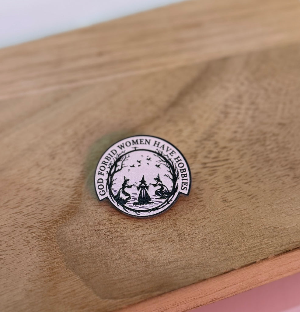 Enamel pin with the phrase "God Forbid Women Have Hobbies" featuring witchy design in black and pink.