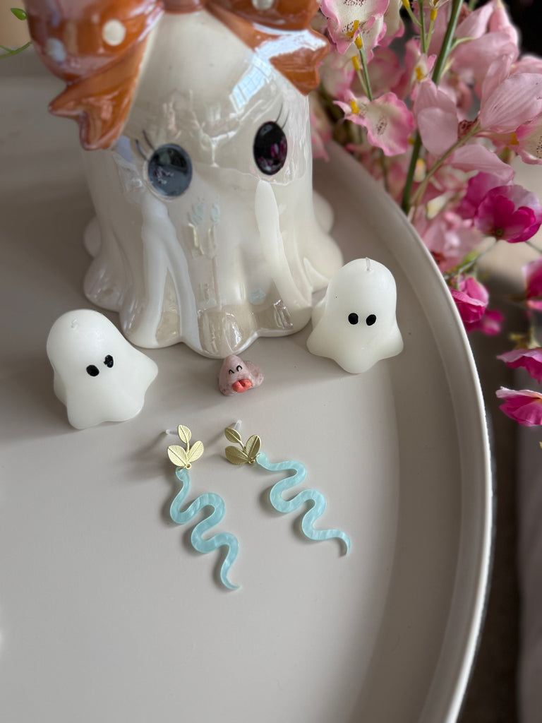 Blue snake-shaped acrylic earrings with gold leaf studs, displayed with ghost candles and flowers.